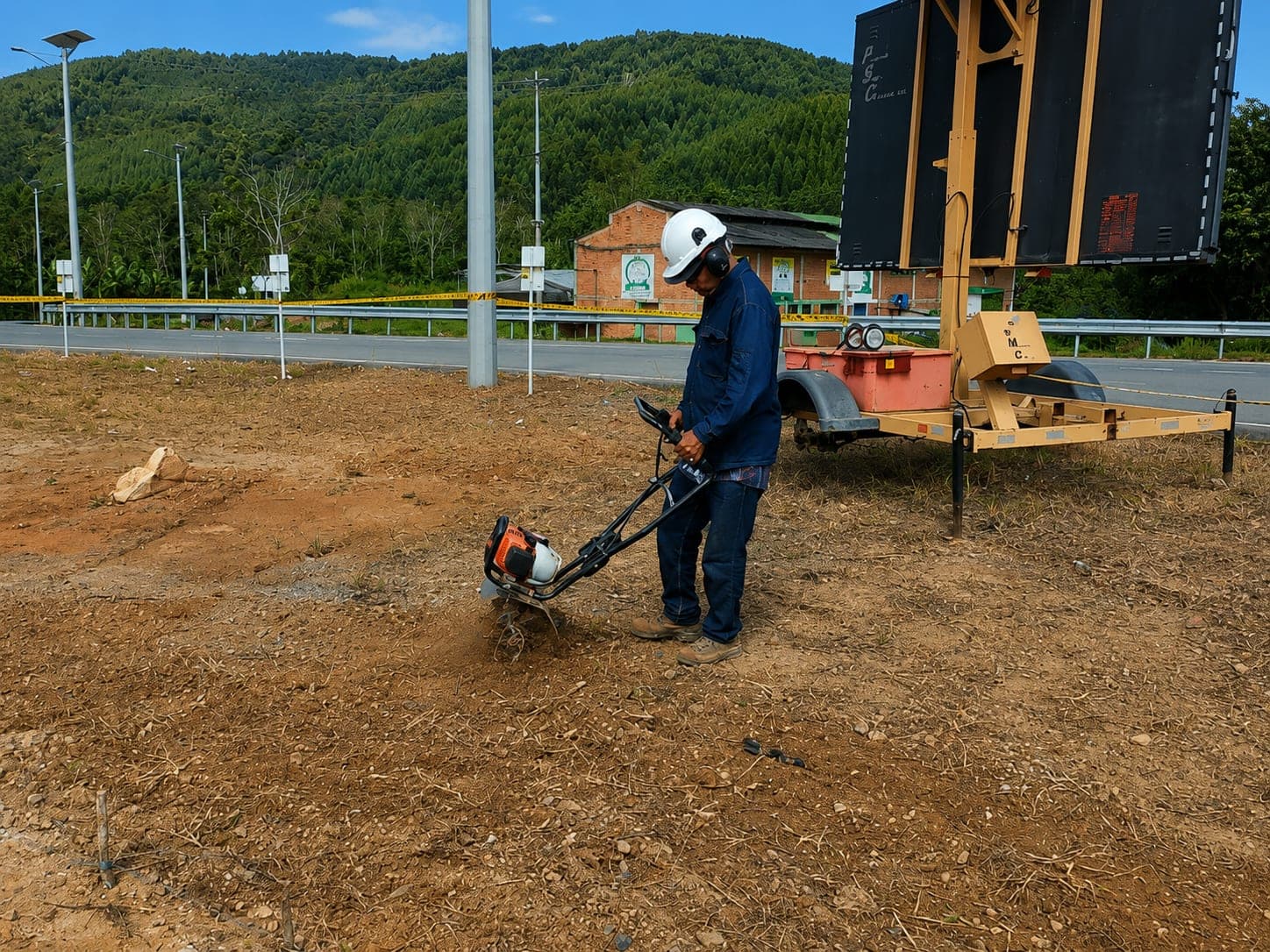 A worker in a helmet operates a mechanical auger on bare ground beside a road, with signage and a trailer in the background.