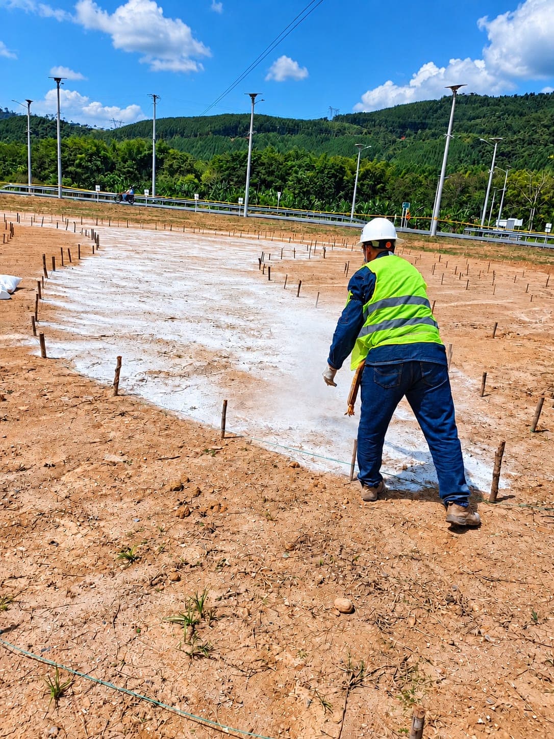 A finished band of stone in the foreground; behind it, two workers prepare the next one among layout stakes.