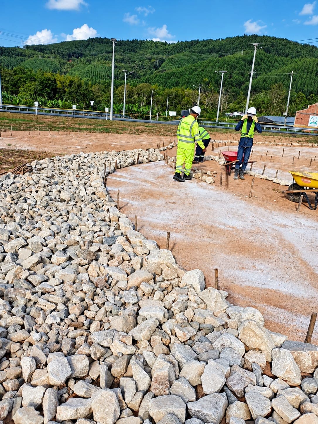 A worker in a helmet loads stone into two wheelbarrows beside a pile, at the edge of a road.