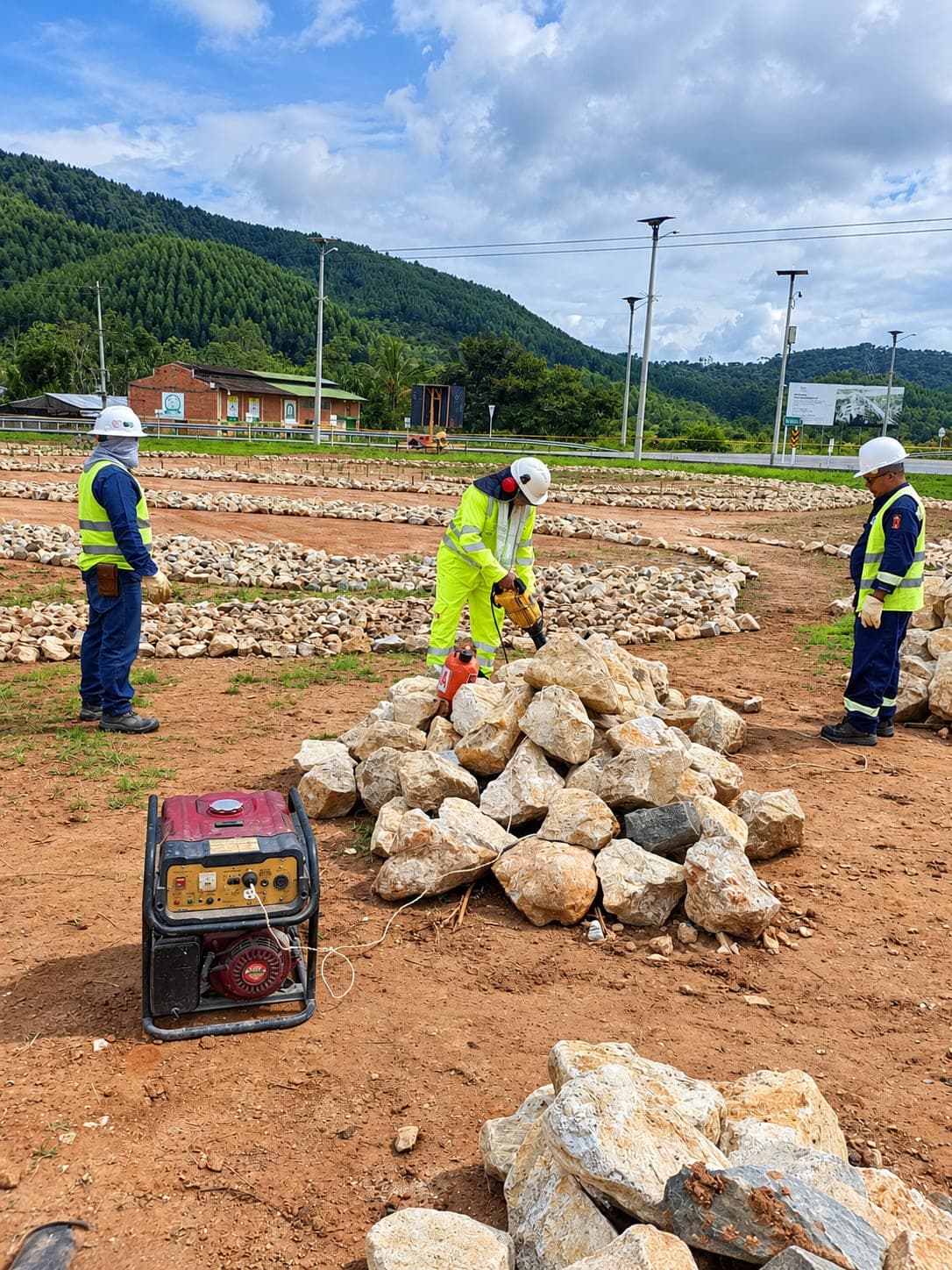 Three workers around a pile of stone; one works a rock with a power tool run off a portable generator.