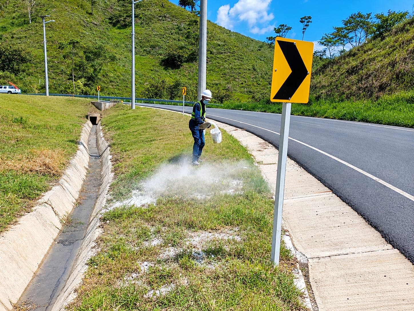 A worker spreads a white soil amendment from a bucket over a road verge, next to a concrete drainage channel.