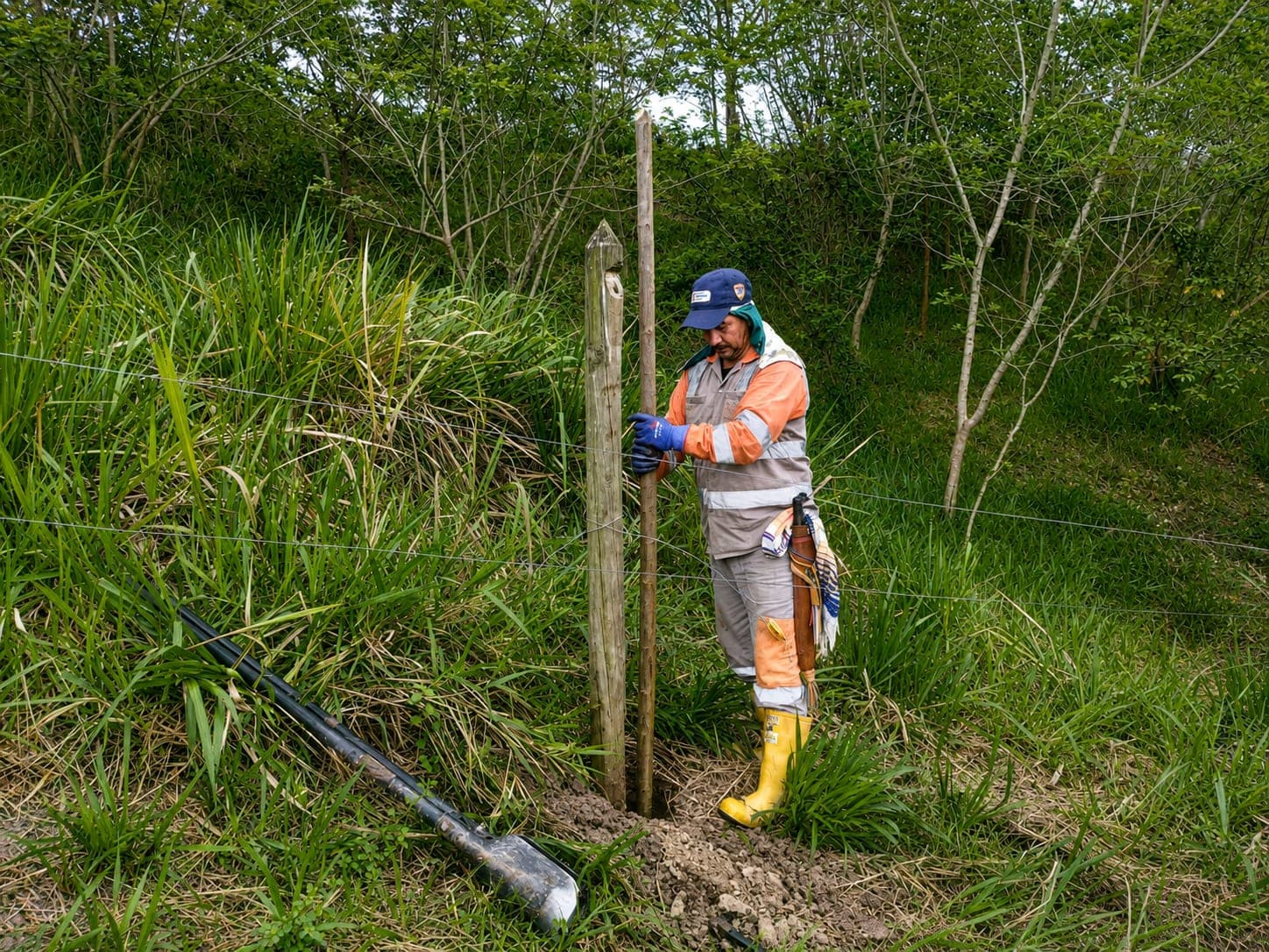 A worker in a helmet and reflective uniform drives in a support stake with a bar beside a young individual on a slope.