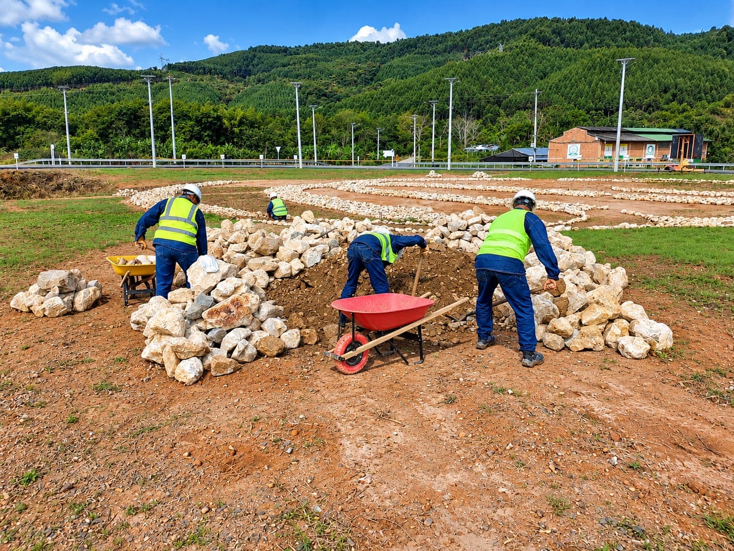 A crew of four lays lines of stone across a flat plot, with wheelbarrows and hills behind.