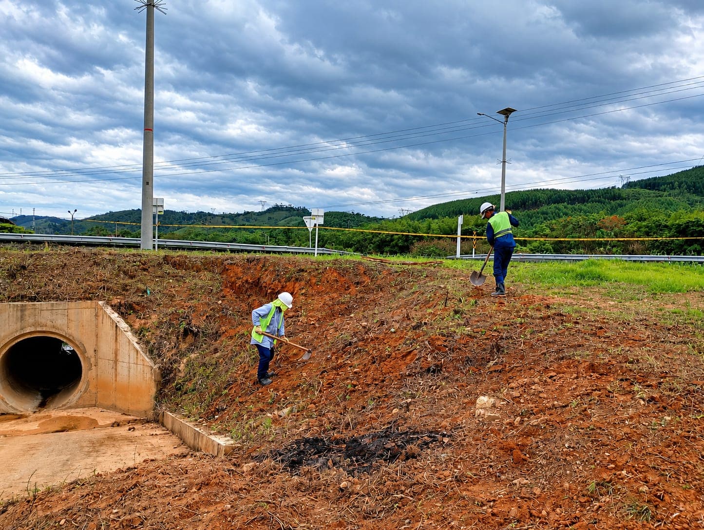 Two workers use hand tools on the earth slope next to a culvert mouth, under a cloudy sky.