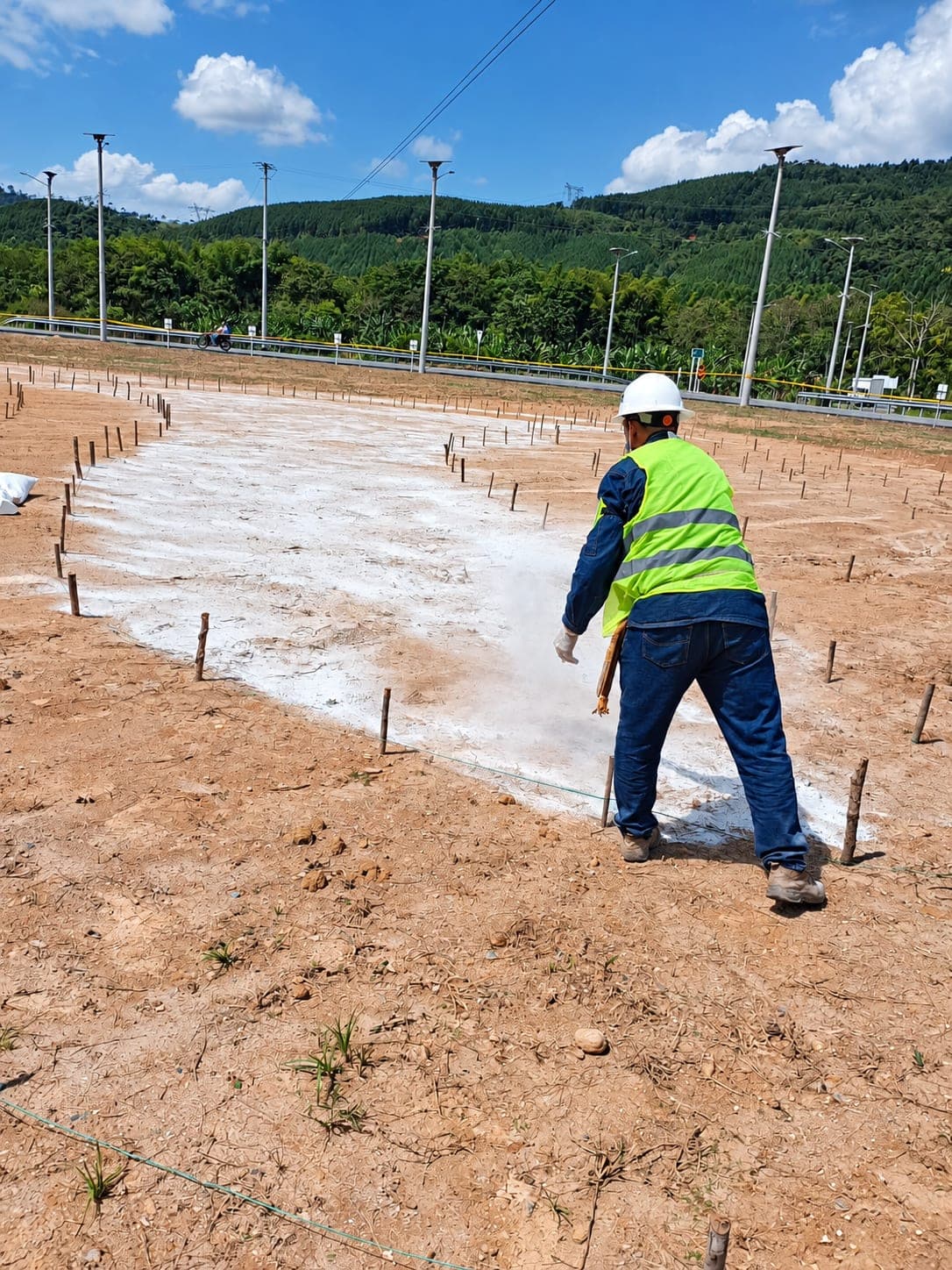 A worker in a helmet and reflective vest marks out a plot with lime, staked with wooden pegs, beside a road.